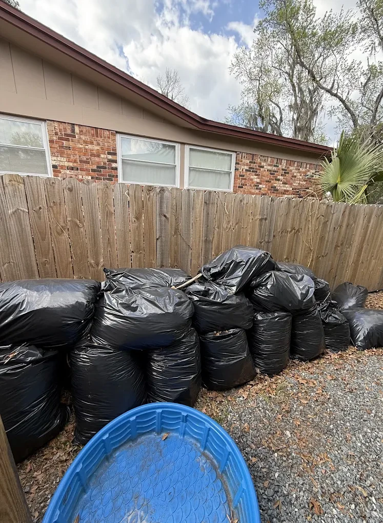 Before photo of bagged yard debris piled along a fence line